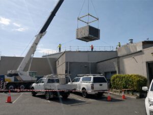 A mobile crane lifting a commercial rooftop package unit into place at a warehouse.