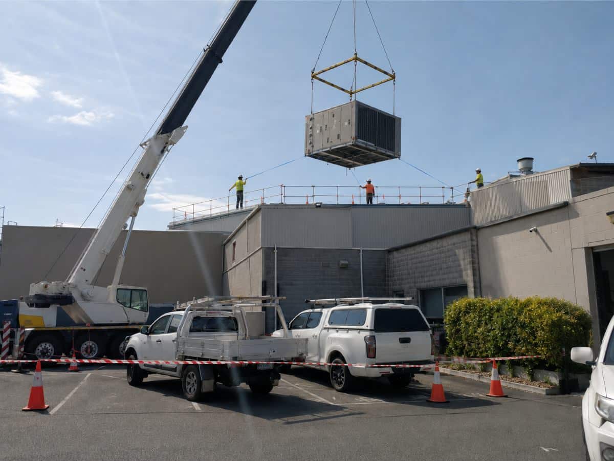 A mobile crane lifting a commercial rooftop package unit into place at a warehouse.
